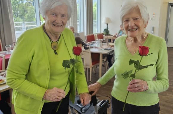 Auf dem Bild sind zwei lächelnde Bewohnerinen zu sehen. Beide tragen grüne Pullover bzw Strickjacke, haben weiße Haare und halten eine Rose in der Hand.