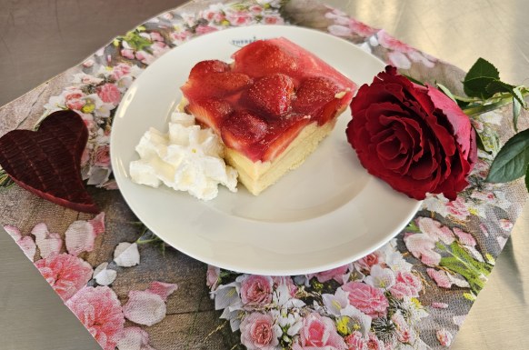 Auf dem Bild ist ein Teller mit selbstgebackenen Erdbeerkuchen und Sahne. Neben dem Teller liegt links ein Holzherz und rechts eine rote Rose.